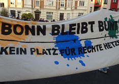 Demonstrierende halten ein großes weißes Transparent, das bunte Farbkleckse aufweist so wie die Botschaft: "BONN BLEIBT BUNT. KEIN PLATZ FÜR RECHTE HETZE."